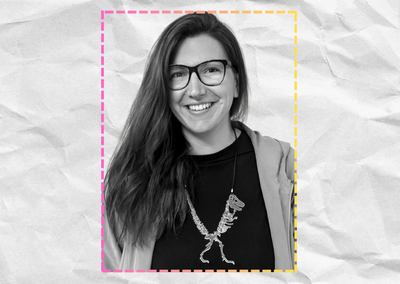 Ilona ist in schwarz-weiß zu sehen, sie schaut in die Kamera, sie trägt eine Brille, ein dunkles Shirt und eine offene, helle Jacke darüber. Um den Hals trägt sie eine Kette, an der ein großes Silbernes Dino-Skelett hängt.